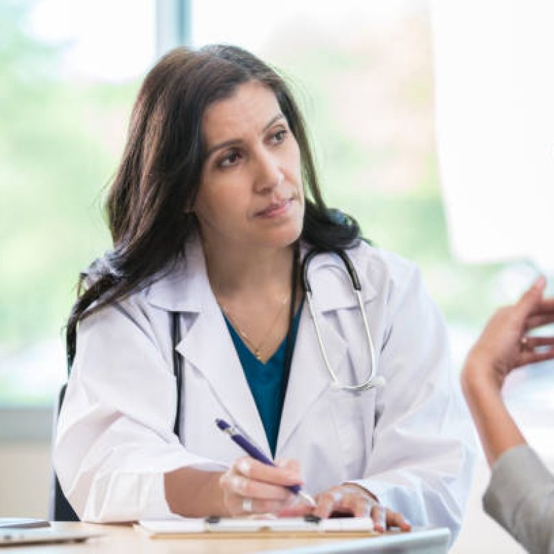 Caring female doctor attentively listens to a young woman talk about her symptoms. The doctor is taking notes notes in the woman's medical chart.